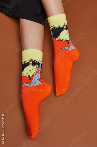 Cropped shot of a girl's foots, lying on a brown background. It is orange artwork socks with abstract man and woman print on foots. 