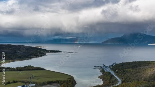 A ferry crossing from Bognes to Lodingen. The fjord is calm, thick white clouds hanging above, a rainbow appears in a distance, HIgh mountains and green fields are framing the scene. photo