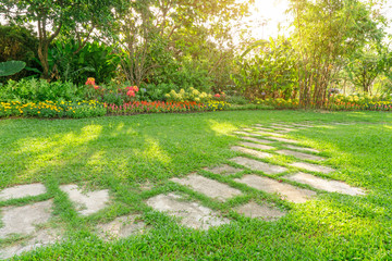 Random pattern of grey concrete stepping stone on green grass lawn, Flowering plant, shrub and trees on background under morning sunlight with good care landscaping in a garden of the public park