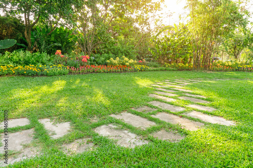Random pattern of grey concrete stepping stone on green grass lawn, Flowering plant, shrub and trees on background under morning sunlight with good care landscaping in a garden of the public park