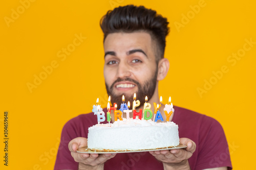 Positive funny young guy with a cap and a burning candle and a homemade cake in his hands posing on a yellow background. Anniversary and birthday concept.