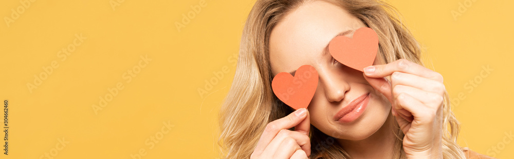 Smiling woman covering eyes with paper heart signs isolated on yellow, panoramic shot Smiling woman covering eyes with paper heart signs isolated on yellow, panoramic shot