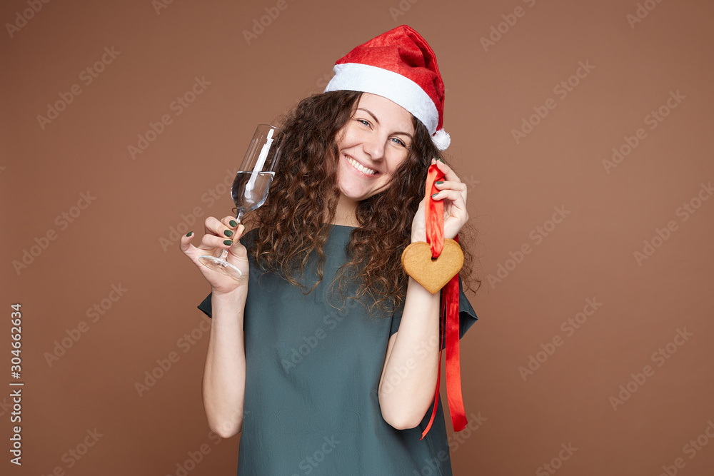 Portrait of happy smiling young woman in green dress and Santa Claus hat standing on brown, looking at camera with ginger cookies and glass of sparkling water, congratulating with New Year's Day. Portrait of happy smiling young woman in green dress and Santa Claus hat standing on brown, looking at camera with ginger cookies and glass of sparkling water, congratulating with New Year's Day.