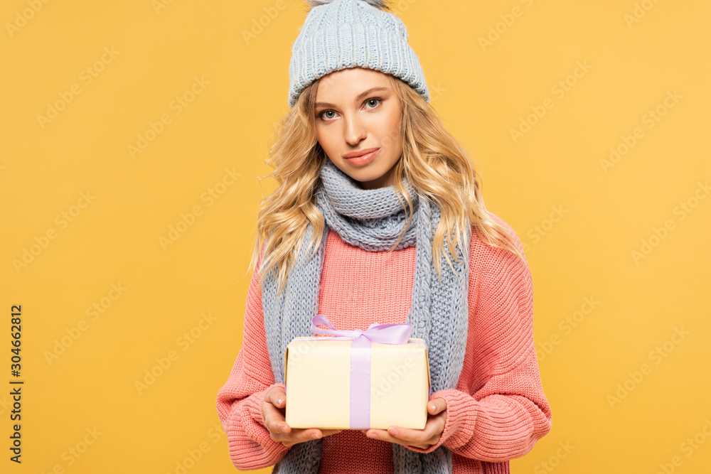 Attractive blonde woman in hat in scarf holding gift box isolated on yellow Attractive blonde woman in hat in scarf holding gift box isolated on yellow