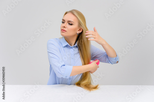 Concept cute model combs the hairstyle sitting at the table. Close-up portrait of a beautiful blonde girl with excellent makeup with long smooth hair on a white background in a blue shirt.