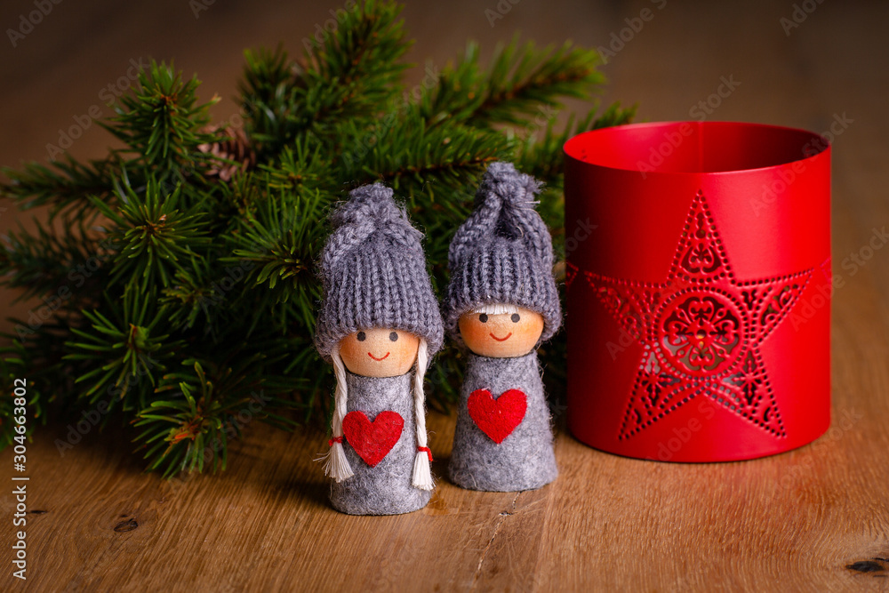 Christmas decoration of wooden table: 2 toy figures (girl, boy) with twig of decorative christmas tree & red table candle holder in the background. Close-up, selective focus. Christmas card.