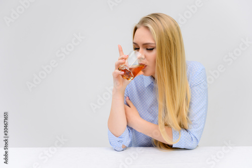 Concept woman with alcohol problems sits at a table with whiskey in a glass. Portrait of a beautiful blonde girl with excellent makeup with long smooth hair on a white background in a blue shirt.