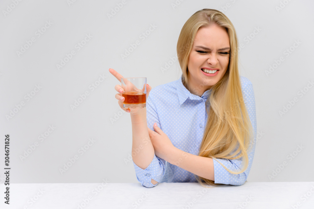 Concept woman with alcohol problems sits at a table with whiskey in a glass. Portrait of a beautiful blonde girl with excellent makeup with long smooth hair on a white background in a blue shirt. Concept woman with alcohol problems sits at a table with whiskey in a glass. Portrait of a beautiful blonde girl with excellent makeup with long smooth hair on a white background in a blue shirt.