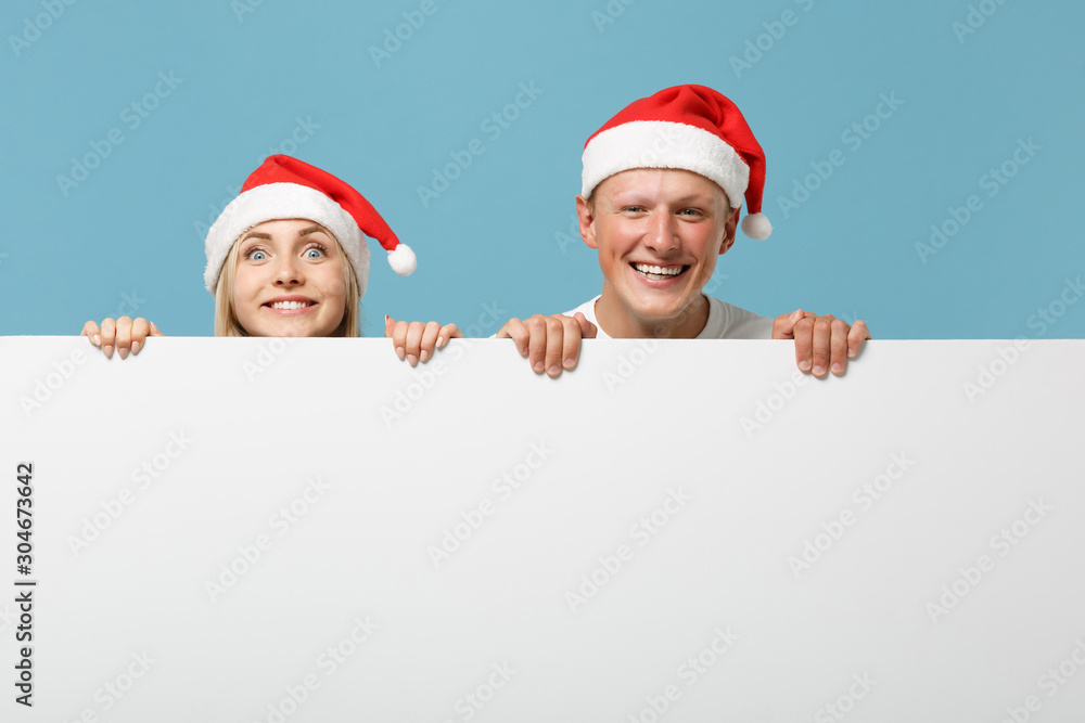 Smiling young Santa couple friends guy girl in Christmas hat isolated on blue background. Happy New Year 2020 celebration holiday concept. Mock up copy space. Holding big white empty blank billboard. Smiling young Santa couple friends guy girl in Christmas hat isolated on blue background. Happy New Year 2020 celebration holiday concept. Mock up copy space. Holding big white empty blank billboard.