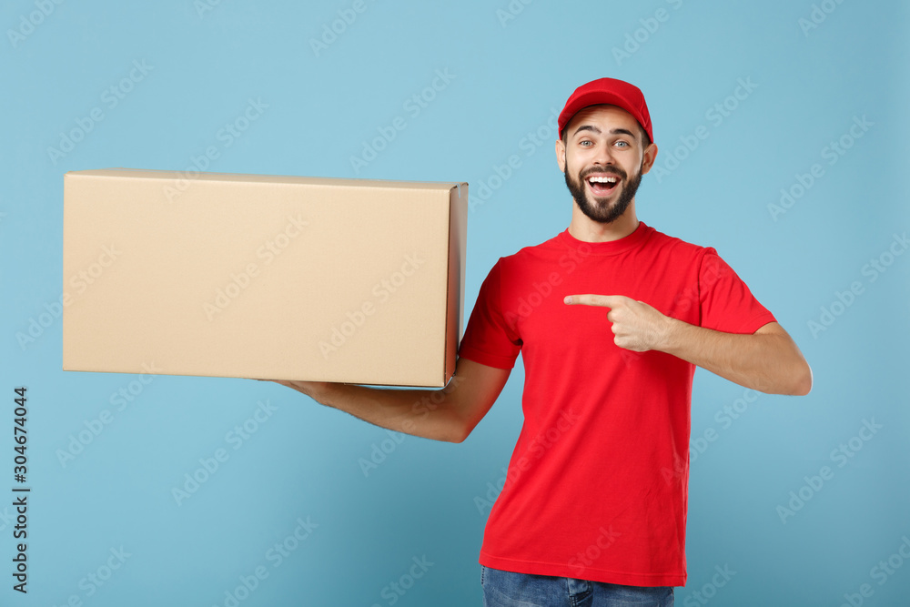 Delivery man in red uniform isolated on blue background, studio portrait. Male employee in cap t-shirt print working as courier dealer hold empty cardboard box. Service concept. Mock up copy space. Delivery man in red uniform isolated on blue background, studio portrait. Male employee in cap t-shirt print working as courier dealer hold empty cardboard box. Service concept. Mock up copy space.