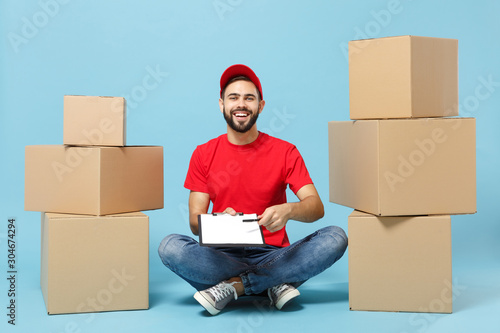 Delivery man in red uniform isolated on blue background, studio portrait. Male employee in cap t-shirt print working as courier dealer sit at empty cardboard box. Service concept. Mock up copy space.