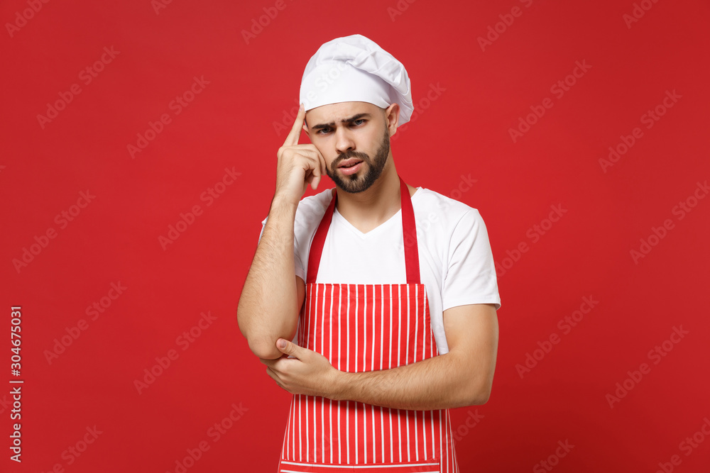 Pensive young bearded male chef cook or baker man in striped apron toque chefs hat posing isolated on red background in studio. Cooking food concept. Mock up copy space. Pointing index finger on head. Pensive young bearded male chef cook or baker man in striped apron toque chefs hat posing isolated on red background in studio. Cooking food concept. Mock up copy space. Pointing index finger on head.