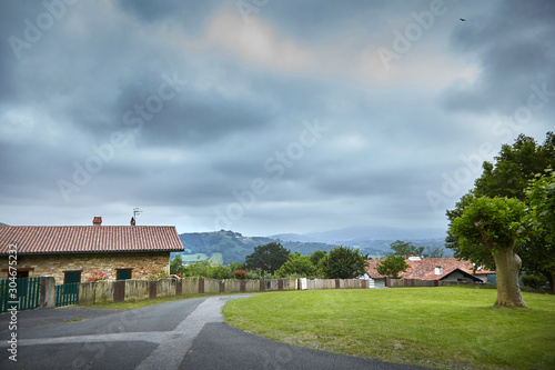 Village, rural landscape at cloudy summer day, farm buildings with tiled roofs. Road, house and fence, countryside, Atlantic Pyrenees, France. Overcast weather