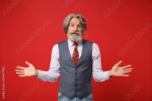 Bewildered confused elderly gray-haired mustache bearded man in classic shirt vest colorful tie isolated on red background in studio. People lifestyle concept. Mock up copy space. Spreading hands. © ViDi Studio