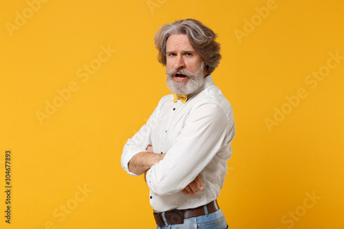 Side view of irritated elderly gray-haired mustache bearded man in white shirt bow tie posing isolated on yellow orange background. People lifestyle concept. Mock up copy space. Holding hands crossed.