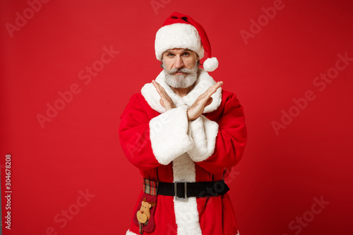 Elderly gray-haired bearded mustache Santa man in Christmas hat posing isolated on red background. Happy New Year 2020 celebration concept. Mock up copy space. Showing stop gesture with crossed hands.