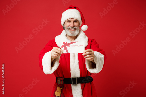 Smiling elderly gray-haired mustache bearded Santa man in Christmas hat isolated on red wall background. Happy New Year 2020 celebration holiday concept. Mock up copy space. Holding gift certificate.
