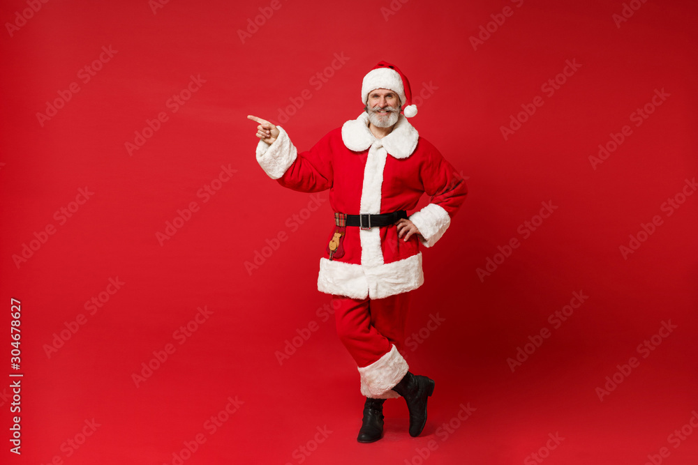 Elderly gray-haired mustache bearded Santa man in Christmas hat posing isolated on red background. Happy New Year 2020 celebration holiday concept. Mock up copy space. Pointing index finger aside. Elderly gray-haired mustache bearded Santa man in Christmas hat posing isolated on red background. Happy New Year 2020 celebration holiday concept. Mock up copy space. Pointing index finger aside.