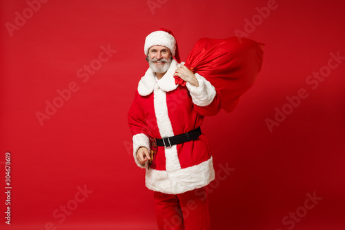 Smiling elderly gray-haired mustache bearded Santa man in Christmas hat posing isolated on red background. Happy New Year 2020 celebration holiday concept. Mock up copy space. Hold bag with presents.