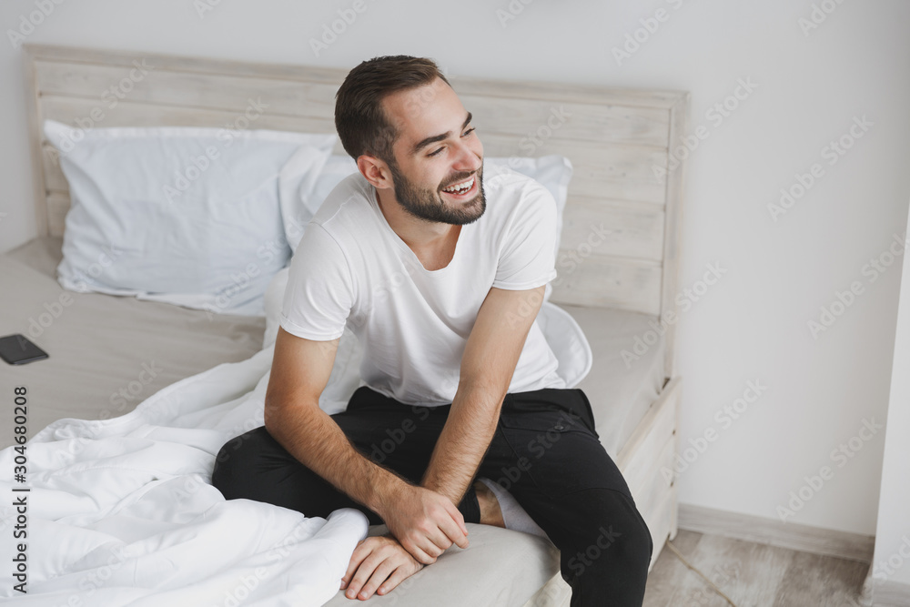 Calm young handsome bearded man sitting in bed with white sheet pillow  blanket in bedroom at