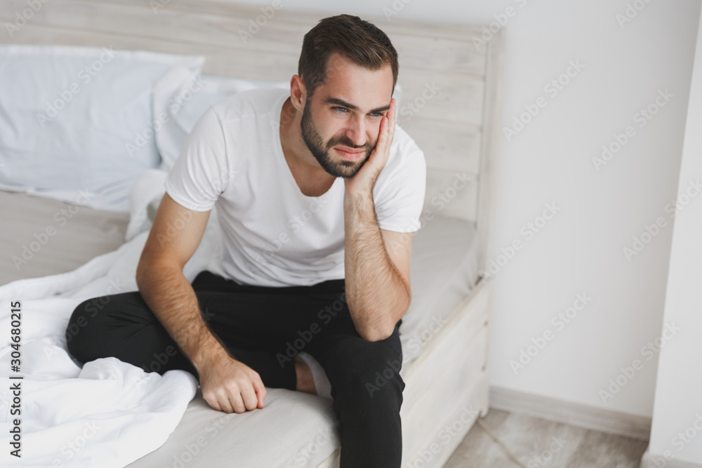 Calm young handsome bearded man sitting in bed with white sheet pillow blanket in bedroom at home. Sad upset male spending time in room. Rest relax good mood lifestyle concept. Mock up copy space. Calm young handsome bearded man sitting in bed with white sheet pillow blanket in bedroom at home. Sad upset male spending time in room. Rest relax good mood lifestyle concept. Mock up copy space.