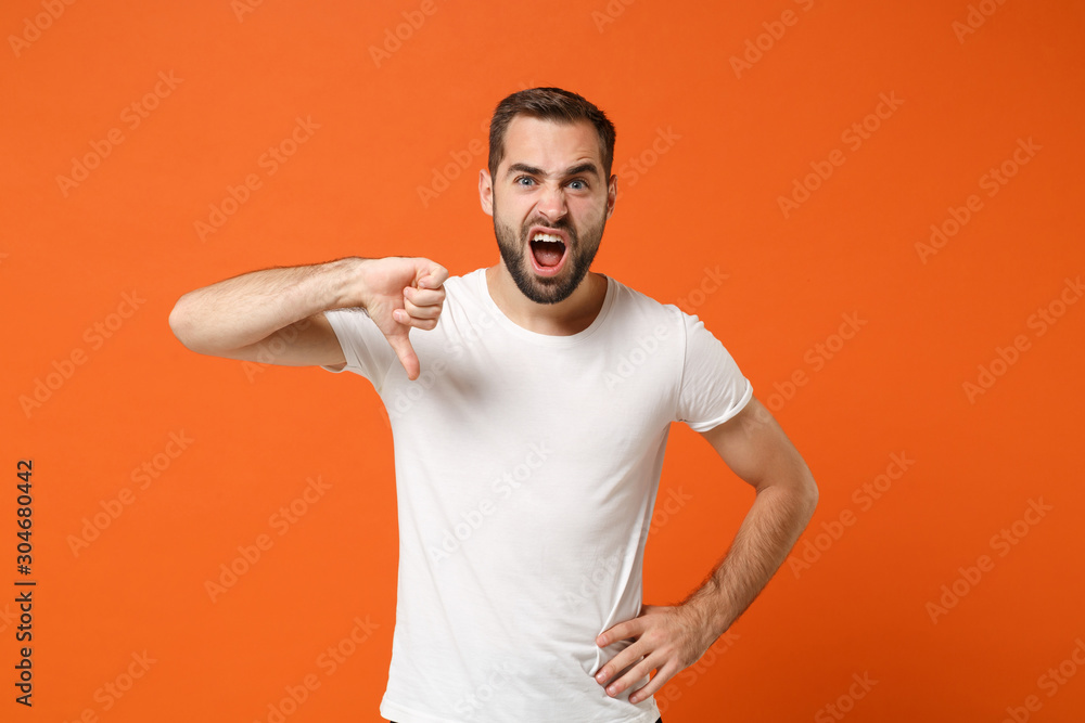 Dissatisfied irritated young man in casual white t-shirt posing isolated on orange wall background studio portrait. People sincere emotions lifestyle concept. Mock up copy space. Showing thumb down. Dissatisfied irritated young man in casual white t-shirt posing isolated on orange wall background studio portrait. People sincere emotions lifestyle concept. Mock up copy space. Showing thumb down.