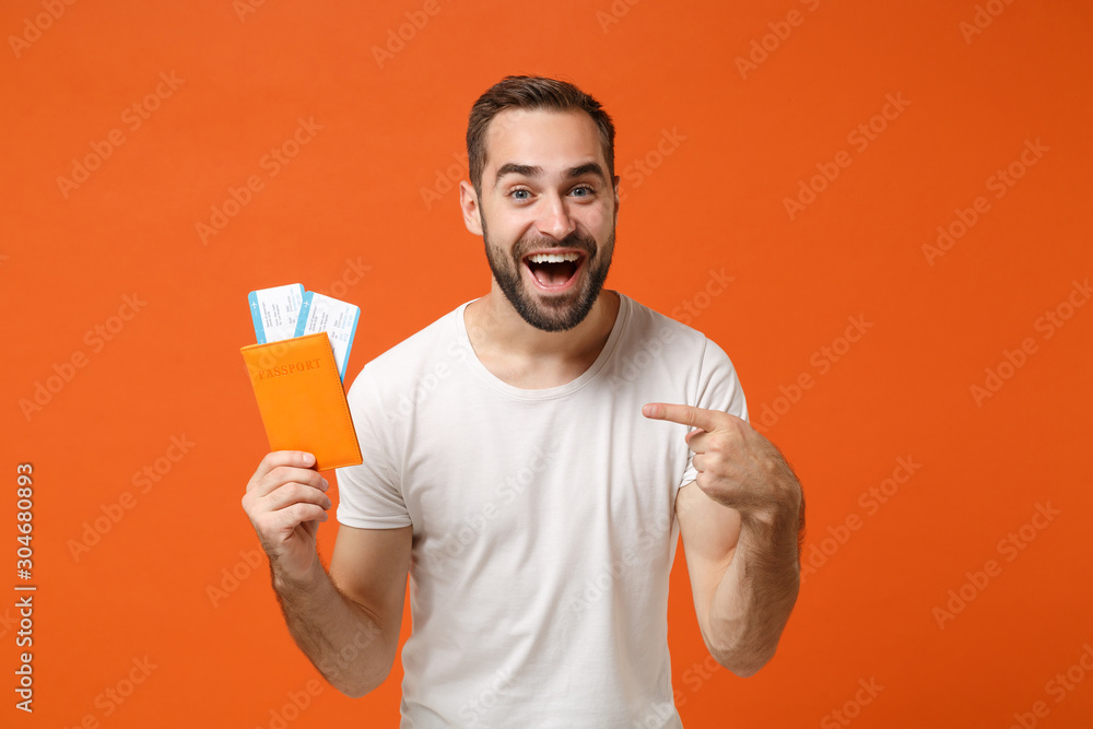 Cheerful young man in casual white t-shirt posing isolated on orange background studio portrait. People lifestyle concept. Mock up copy space. Pointing index finger on passport boarding pass ticket. - obrazy, fototapety, plakaty 