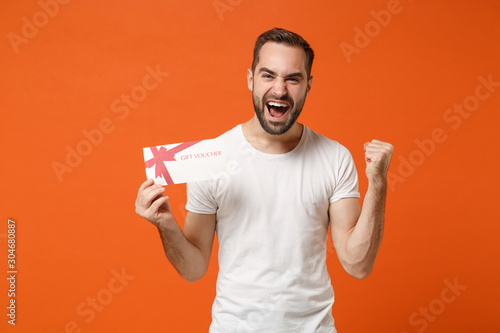 Overjoyed young man in casual white t-shirt posing isolated on orange background in studio. People sincere emotions lifestyle concept. Mock up copy space. Hold gift certificate, doing winner gesture. © ViDi Studio