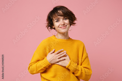 Smiling pleasant young brunette woman girl in yellow sweater posing isolated on pastel pink background, studio portrait. People lifestyle concept. Mock up copy space. Holding hands crossed on heart.