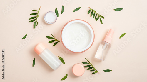 Organic cosmetic bottle containers and jar with white face cream on beige background with green herbal leaves. Creative layout, minimal flat lay style composition. Natural beauty product concept.