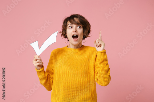 Shocked young brunette woman girl in yellow sweater posing isolated on pastel pink background. People lifestyle concept. Mock up copy space. Hold check mark, point index finger up with great new idea.