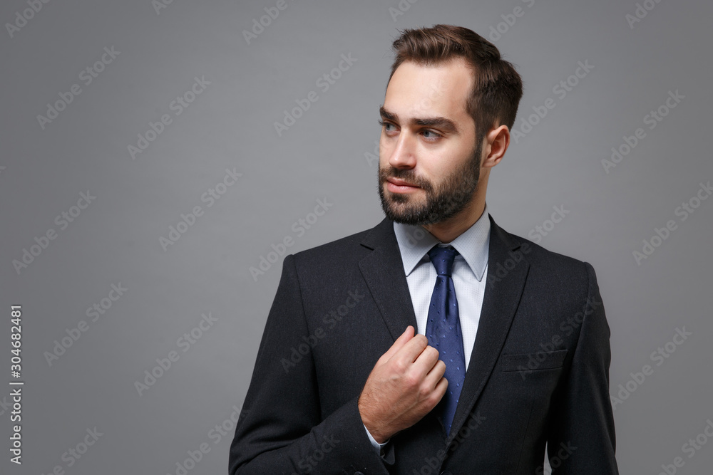 Confident young bearded business man in classic black suit shirt tie posing isolated on grey background studio portrait. Achievement career wealth business concept. Mock up copy space. Looking aside. Confident young bearded business man in classic black suit shirt tie posing isolated on grey background studio portrait. Achievement career wealth business concept. Mock up copy space. Looking aside.