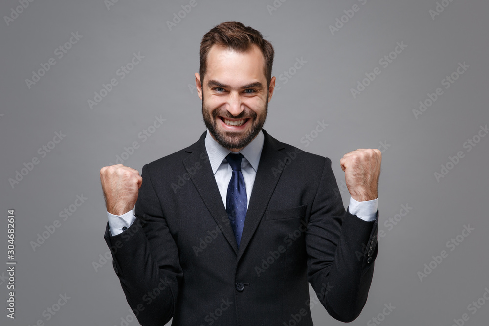 Smiling young business man in classic black suit shirt tie posing isolated on grey background in studio. Achievement career wealth business concept. Mock up copy space. Clenching fists like winner. Smiling young business man in classic black suit shirt tie posing isolated on grey background in studio. Achievement career wealth business concept. Mock up copy space. Clenching fists like winner.