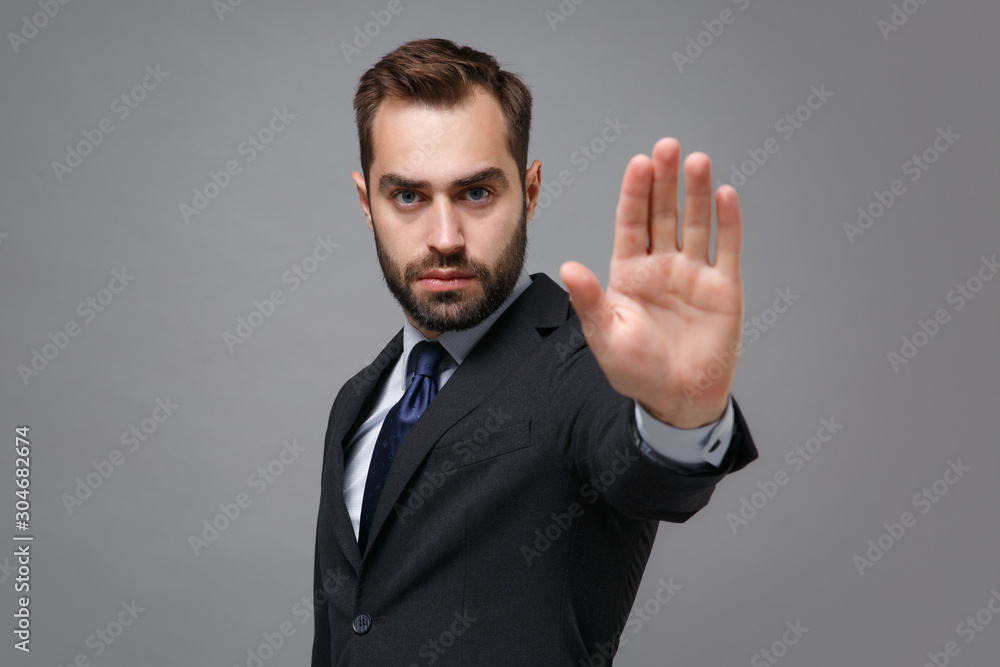 Serious young bearded business man in classic black suit shirt tie posing isolated on grey background. Achievement career wealth business concept. Mock up copy space. Showing stop gesture with palm. - obrazy, fototapety, plakaty 