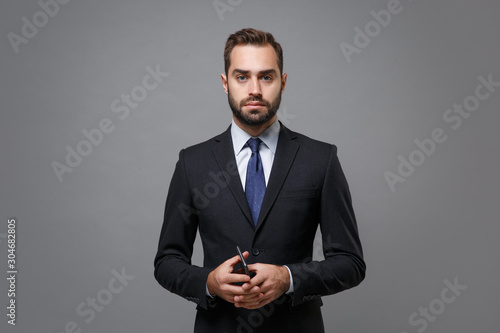 Serious young bearded business man in classic suit shirt tie posing isolated on grey background studio portrait. Achievement career wealth business concept. Mock up copy space. Holding mobile phone.