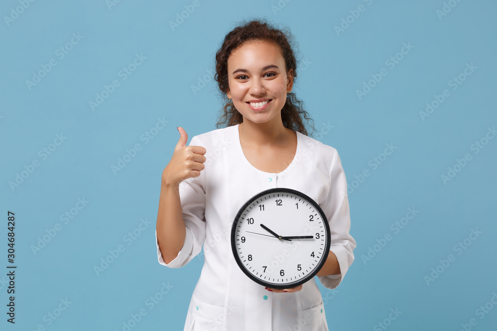 Smiling african american female doctor woman in white medical gown hold clock showing thumb up isolated on blue background in studio. Healthcare personnel medicine health concept. Mock up copy space. Smiling african american female doctor woman in white medical gown hold clock showing thumb up isolated on blue background in studio. Healthcare personnel medicine health concept. Mock up copy space.