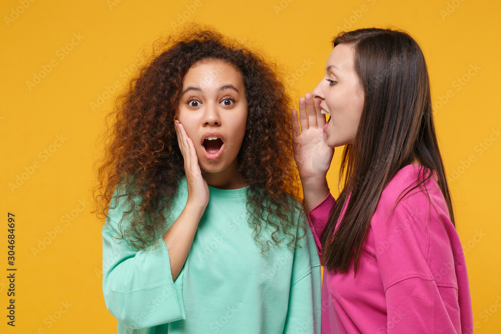 Two shocked women friends european african american in pink green clothes isolated on yellow background. People lifestyle concept. Mock up copy space. Whispering secret behind hand, put palm on cheek. Two shocked women friends european african american in pink green clothes isolated on yellow background. People lifestyle concept. Mock up copy space. Whispering secret behind hand, put palm on cheek.