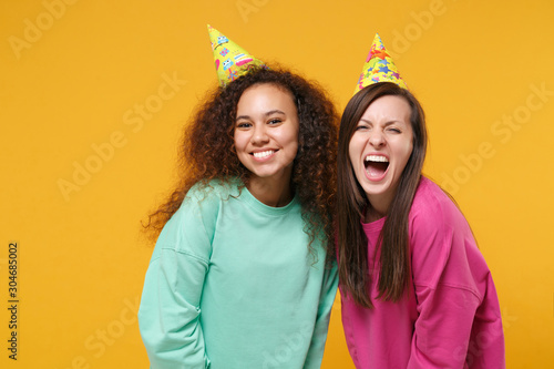 Two smiling screaming women friends european and african american girls in pink green clothes, birthday hats posing isolated on yellow orange background. People lifestyle concept. Mock up copy space.