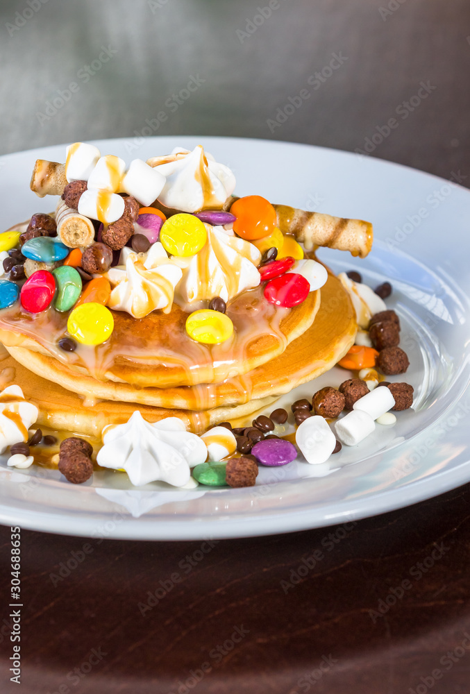 Pancakes in a white plate are decorated with colorful candies, marshmallows and topped with caramel topping Pancakes in a white plate are decorated with colorful candies, marshmallows and topped with caramel topping