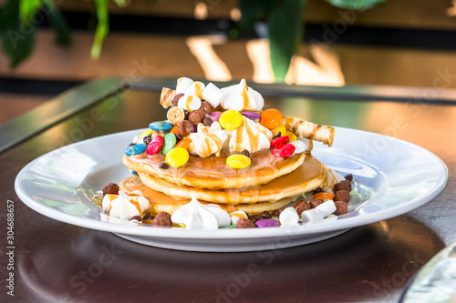 Pancakes in a white plate are decorated with colorful candies, marshmallows and topped with caramel topping