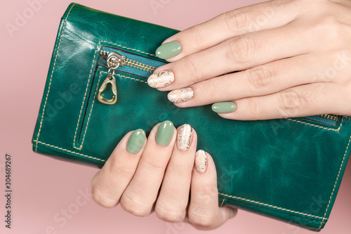 Beautiful white woman holding her green leather wallet isolated at pink pastel background. Close up view of trendy stylish modern manicure of green and beige colors. Horizontal color photography.
