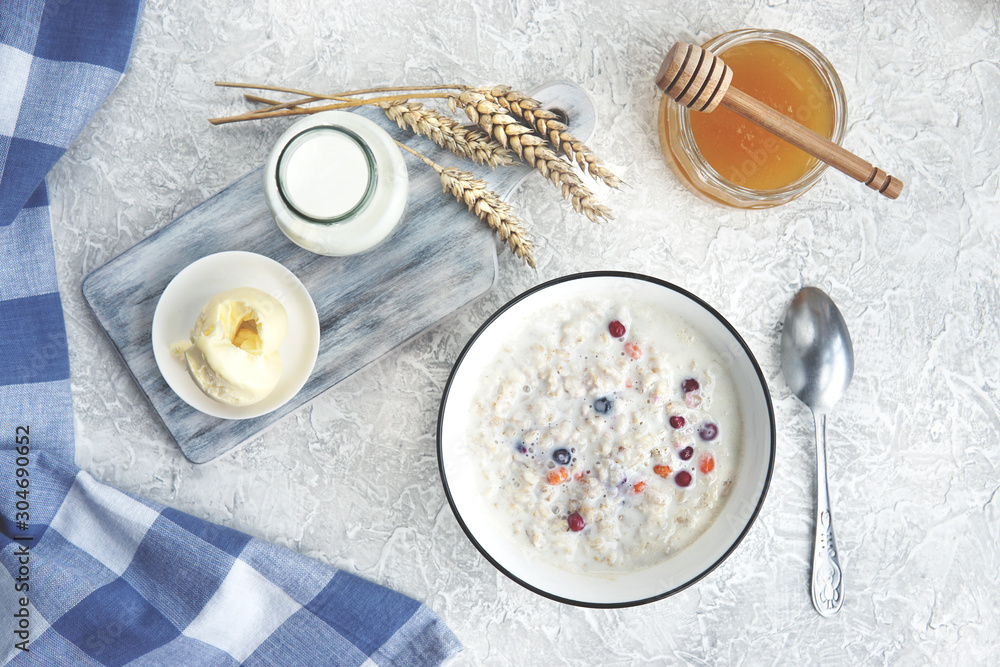 Bowl of oatmeal porridge with berries, honey, bottle of milk, butter, wooden rustic board, grey table near window, hot and healthy food for Breakfast, top view, flat lay. Bowl of oatmeal porridge with berries, honey, bottle of milk, butter, wooden rustic board, grey table near window, hot and healthy food for Breakfast, top view, flat lay.