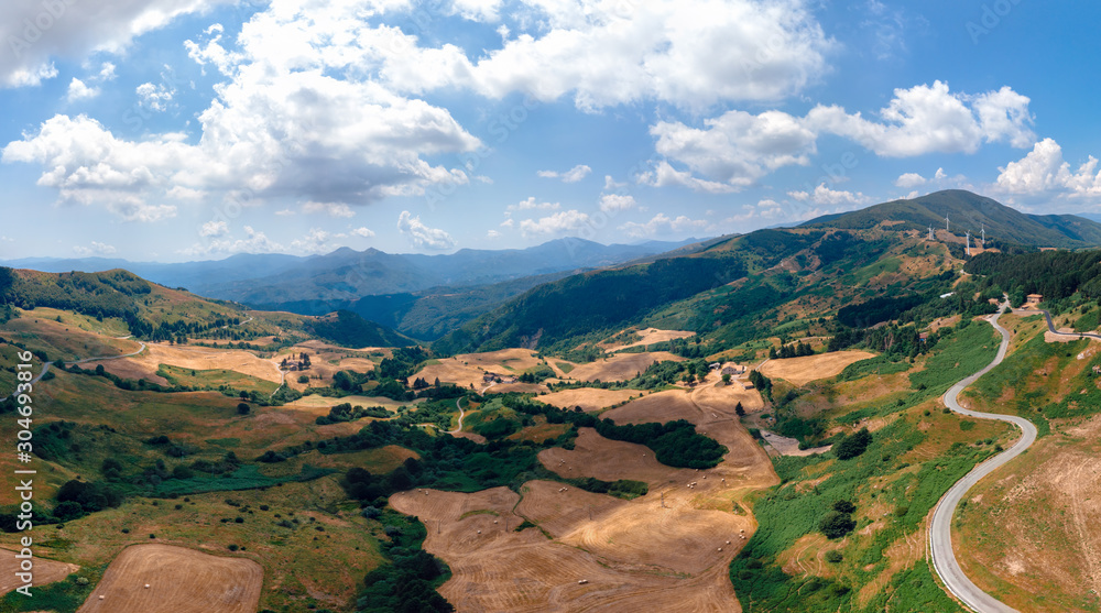 Fototapeta premium Aerial wide view panorama of fields and winding roads, farms, harvested grass, haystacks. Wind turbines in mountain, Passo Di Cento, Taglieto, Varese Ligure, Province of La Spezia, Italy