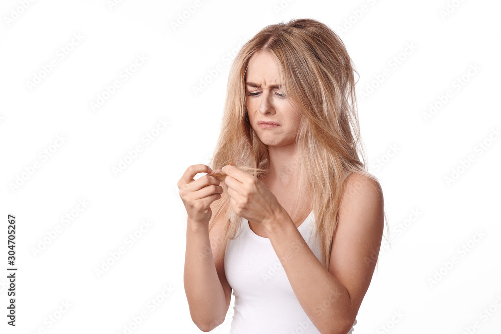 Pretty young woman having a bad hair day standing looking dejectedly at the dry ends of her tousled long blond hair as she holds them up in her hands isolated on white Pretty young woman having a bad hair day standing looking dejectedly at the dry ends of her tousled long blond hair as she holds them up in her hands isolated on white
