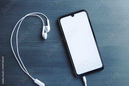 Top view of Mobile phone with white screen and headphones on the table.