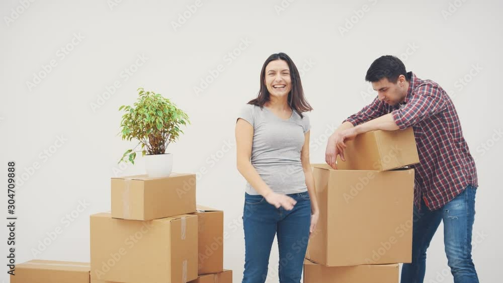 Young man is totaly exhausted. He can not carry heavy boxes from one side to another. Woman is standing, smiling, saying hyrray, pointing where to put boxes. Young man is totaly exhausted. He can not carry heavy boxes from one side to another. Woman is standing, smiling, saying hyrray, pointing where to put boxes.