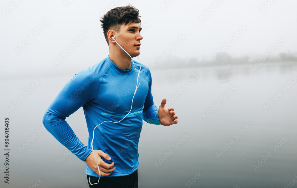 Horizontal outdoor image of young runner man running in the morning on the fog lake background. Fitness male exercising in the park and listenting the music on earphones. People and sport concept - obrazy, fototapety, plakaty 