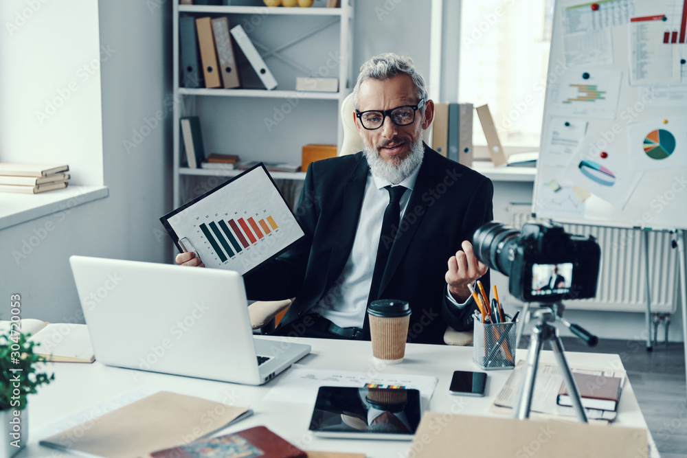 Confident mature man in elegant suit showing chart and sharing business experience while making social media video Confident mature man in elegant suit showing chart and sharing business experience while making social media video