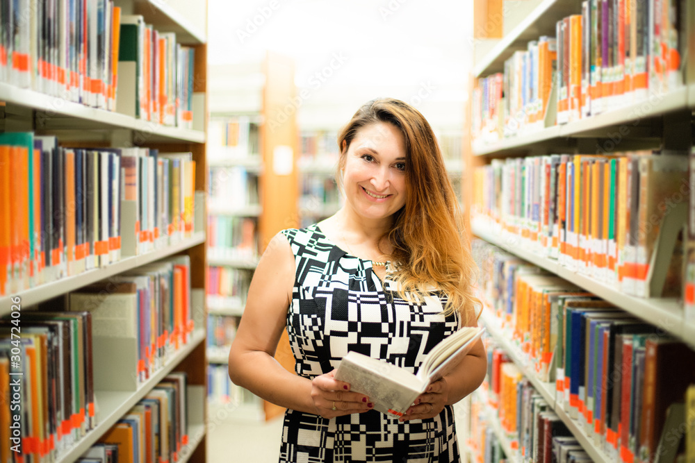 Beautiful white woman female student smiling and looking on the camera and holding book. She is standing near, between bookshelves in modern interior library of university Beautiful white woman female student smiling and looking on the camera and holding book. She is standing near, between bookshelves in modern interior library of university