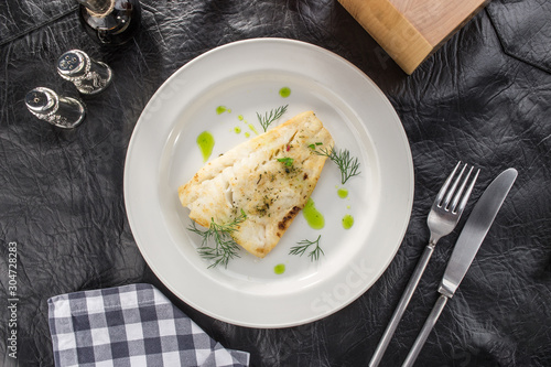fried white fish fillet on white plate with tomato juice on black leather table